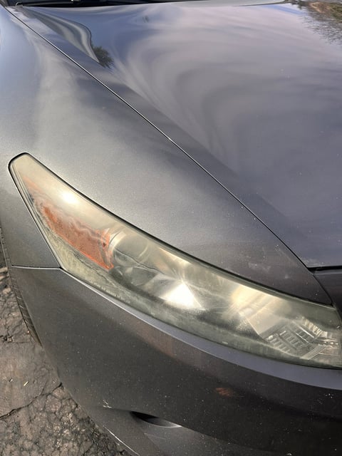 Close-up of a silver cars headlight with oxidized lens and weathered hood in daylight